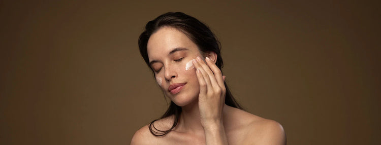 Woman applying Antipodes moisturiser to her cheek against a brown gradient background.