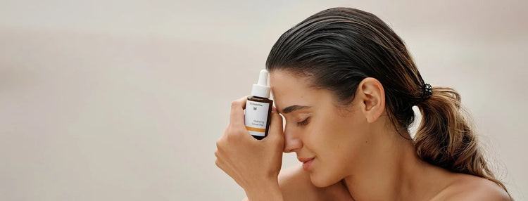 Woman holding a dropper bottle of Dr. Hauschka's Hydrating Serum Plus against her nose and looking glowy and beautiful with a neutral background.