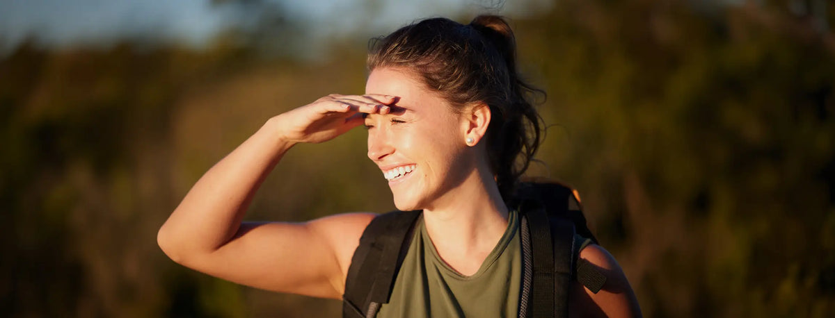 A woman hiking outdoors in a forest, wearing an olive green tank top and a black backpack, with her hand shielding her eyes from the sun.