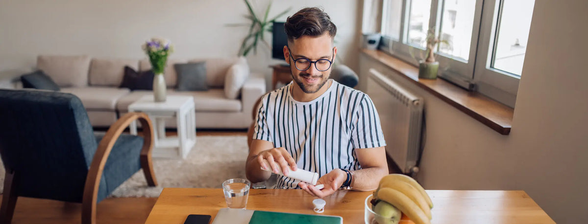 A man wearing glasses and a blue-and-white striped t-shirt sits at a wooden table in a bright living room, holding a supplement bottle. A banana, a glass of water, and a green notebook are on the table in front of him.