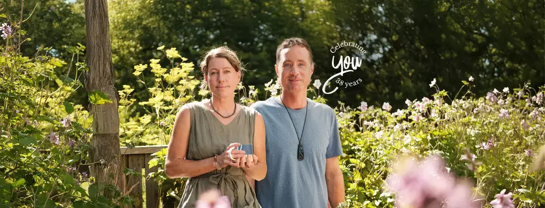 A man and a woman standing together in a sunlit garden with green foliage and flowers in the background.