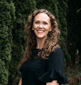 Woman with long curly hair wearing a black top standing in front of greenery.