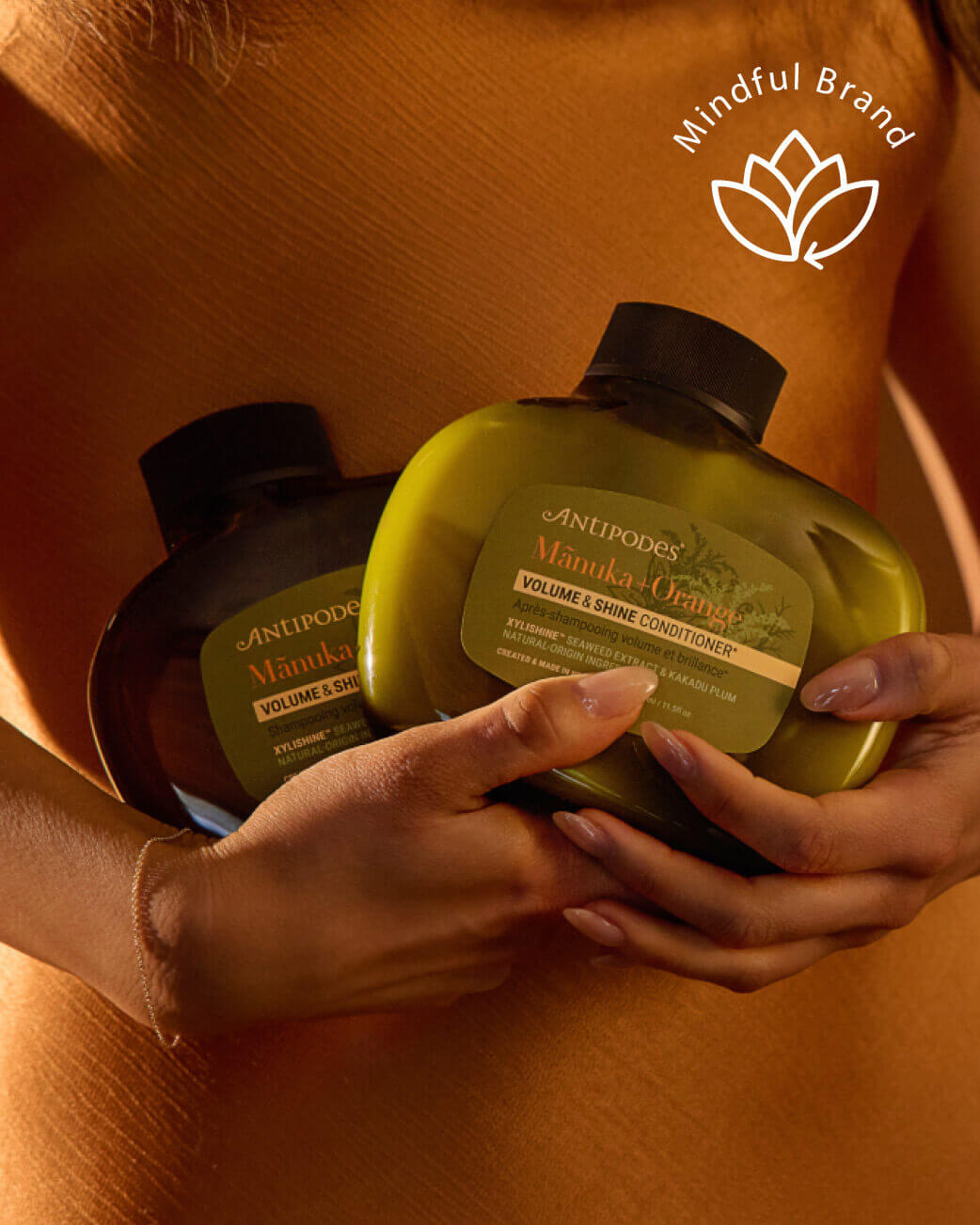 Two bottles of Antipodes hair care products held by hands against an orange background, with 'Mindful Brand' logo.