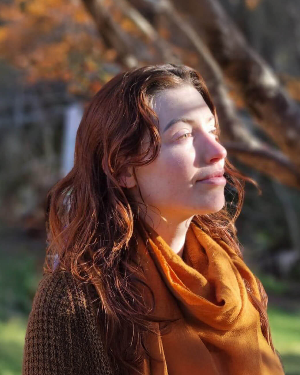 Woman wearing an orange scarf and brown sweater in a natural setting with trees and grass.