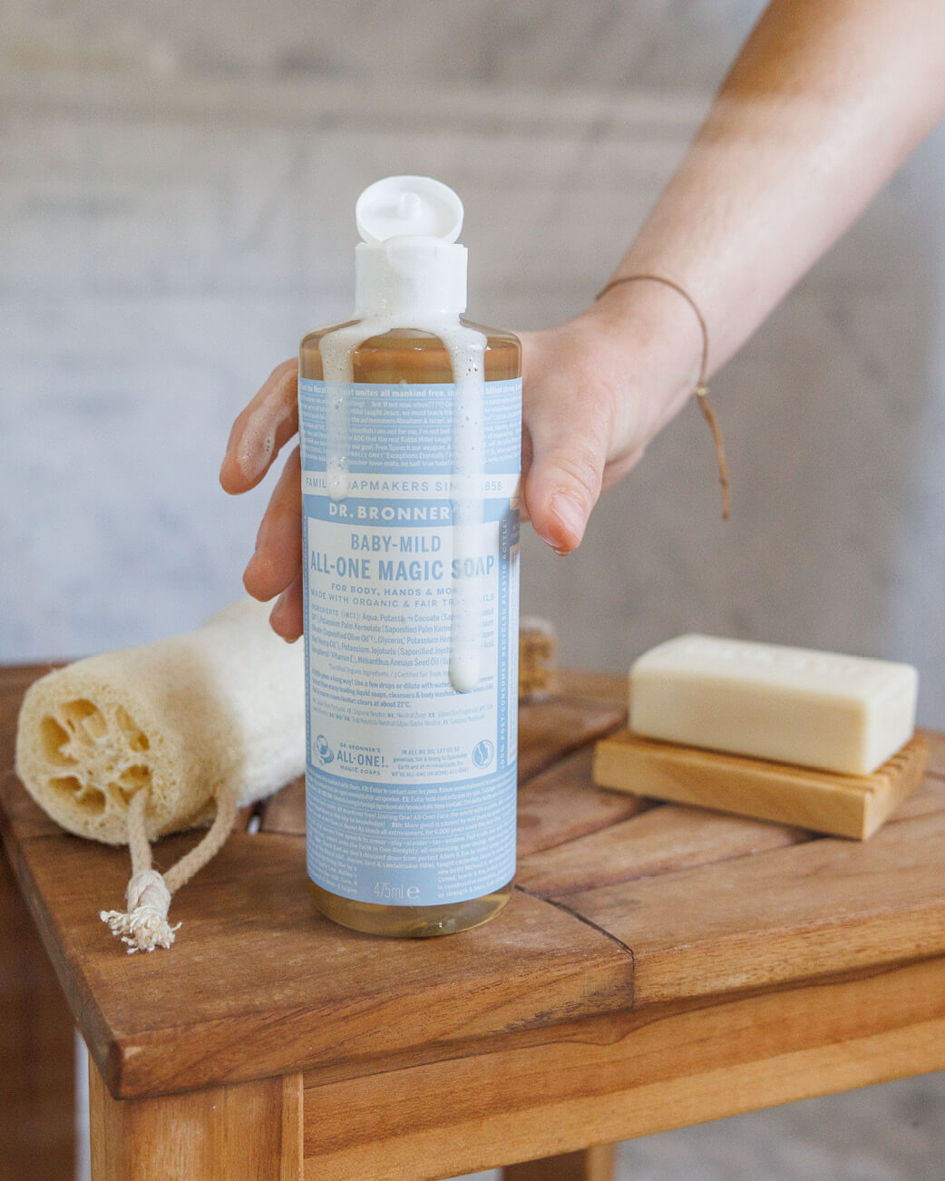 Person holding a bottle of Dr. Bronner's Baby-Mild All-One Magic Soap on a wooden surface.
