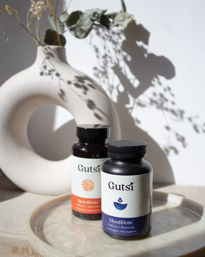 Two Gutsi supplement bottles on a textured surface with a decorative vase and plants in the background.