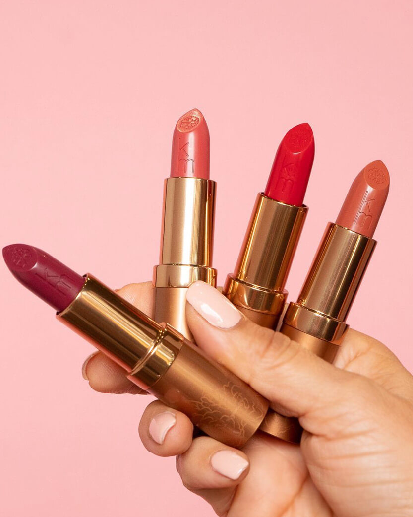 Four lipsticks held in a hand against a pink background.