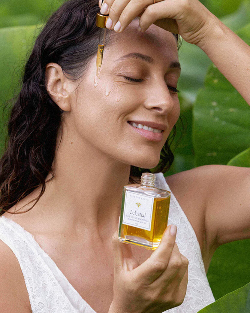 Woman applying a droplet of oil to her face in a natural setting.