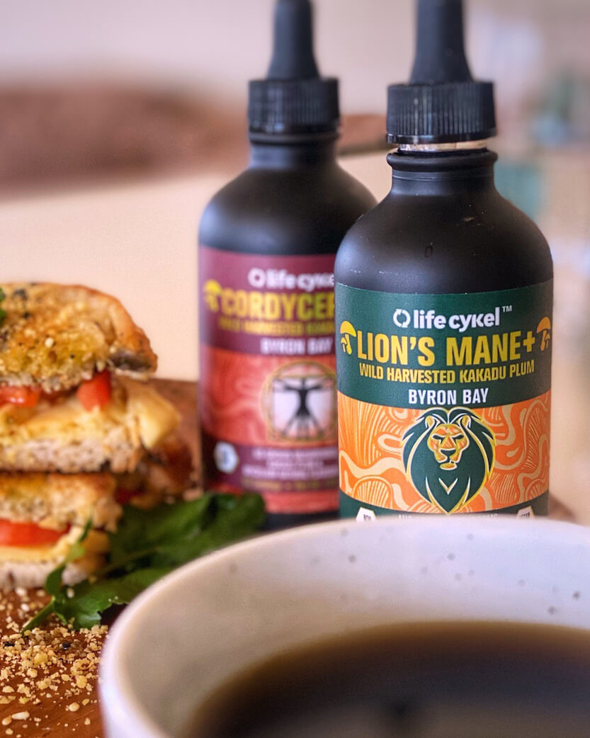 Two bottles of Life Cykel mushroom products with a sandwich and coffee in the background.