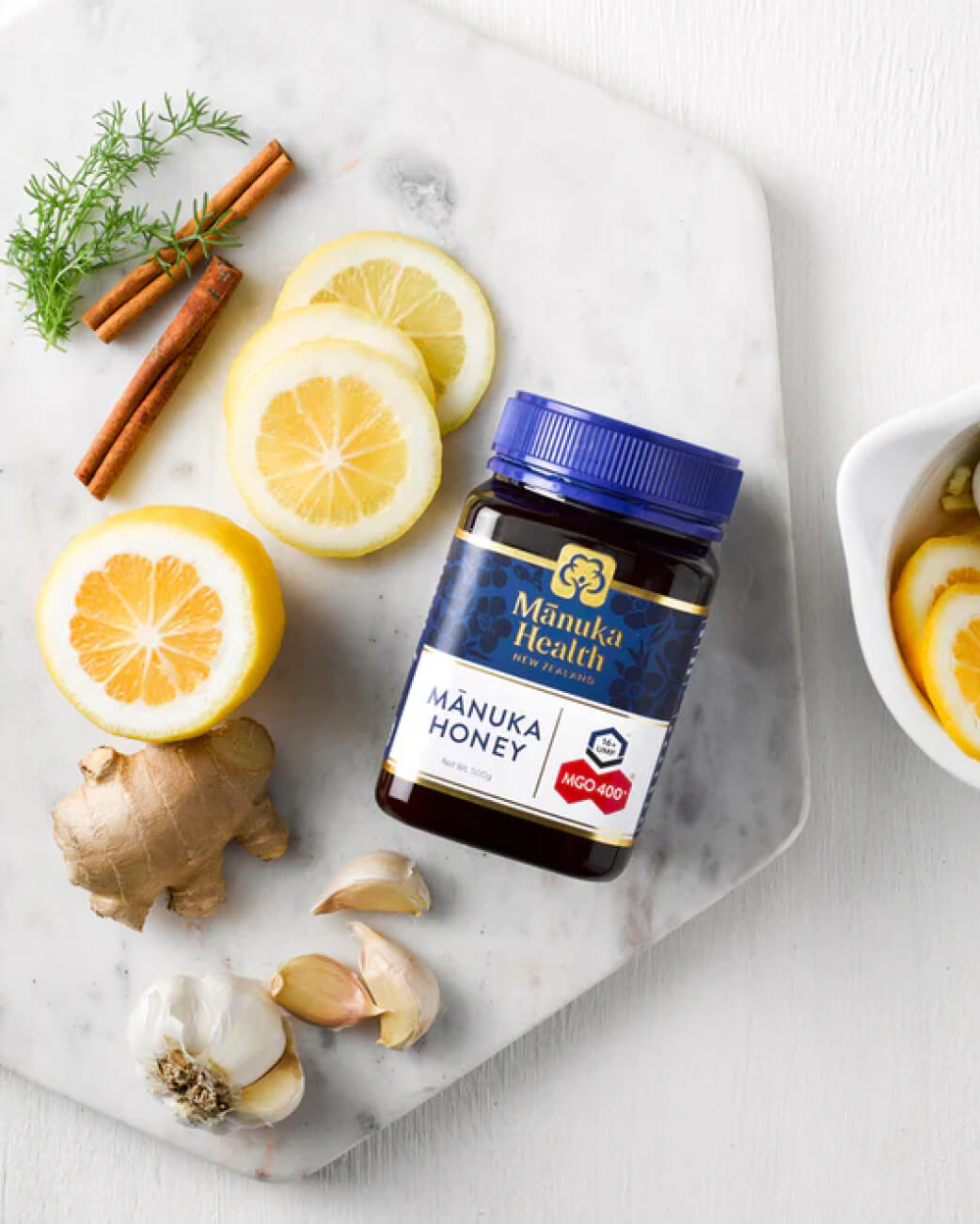 Manuka Health honey jar on a marble slab with lemons, ginger, and cinnamon sticks.