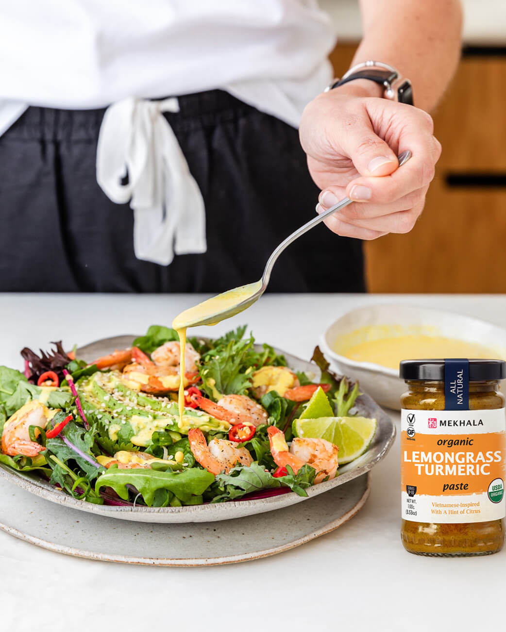 Person pouring sauce over a salad with a bottle of Mekhala Lemongrass Turmeric paste on a table.