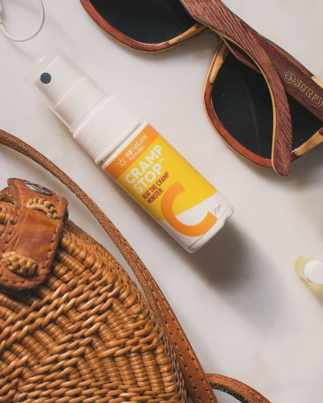 Cramp Stop bottle with sunglasses and a woven bag on a light surface.