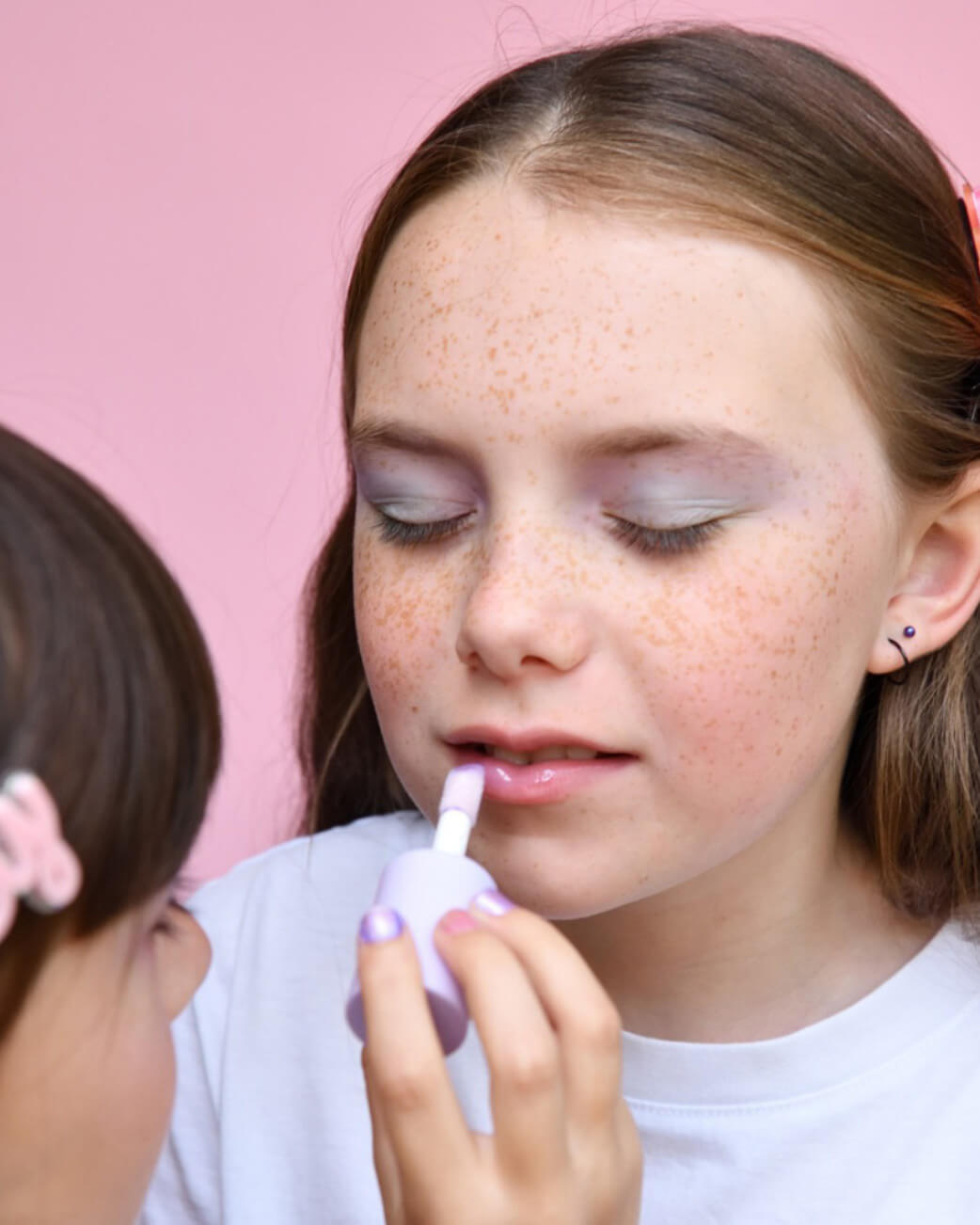 Person applying lip gloss to another person's lips with a pink background.