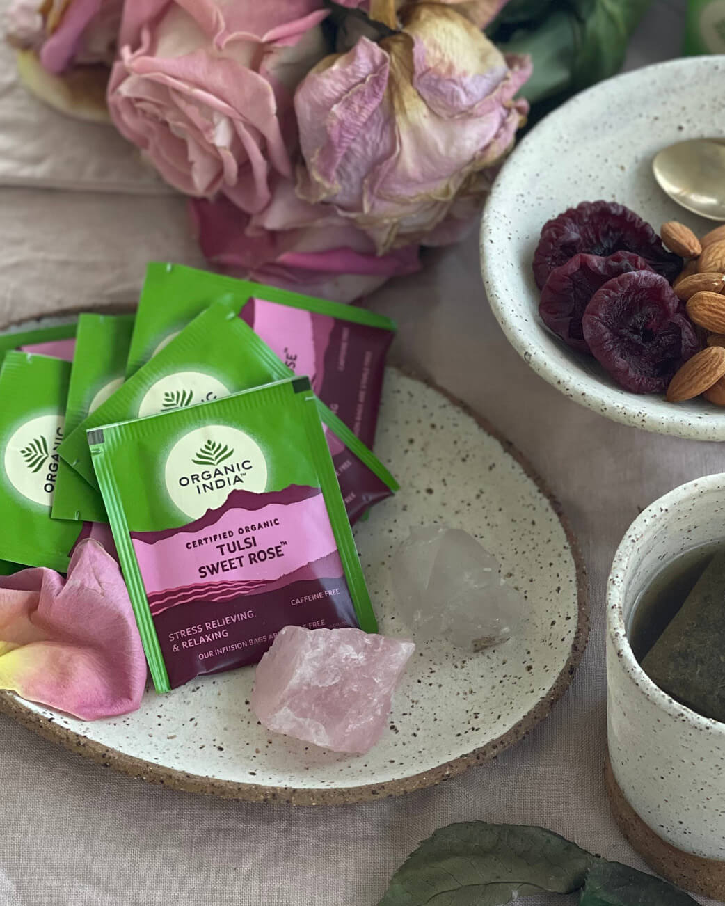 Organic India tulsi sweet rose tea packets on a plate with flowers and a bowl of dried fruits.
