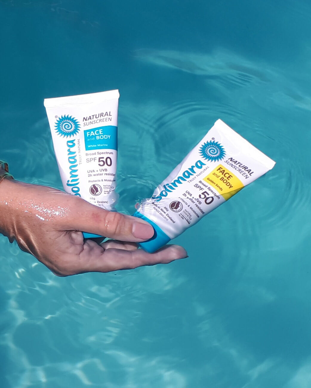 Two tubes of Solimara Natural Sunscreen being held over water.