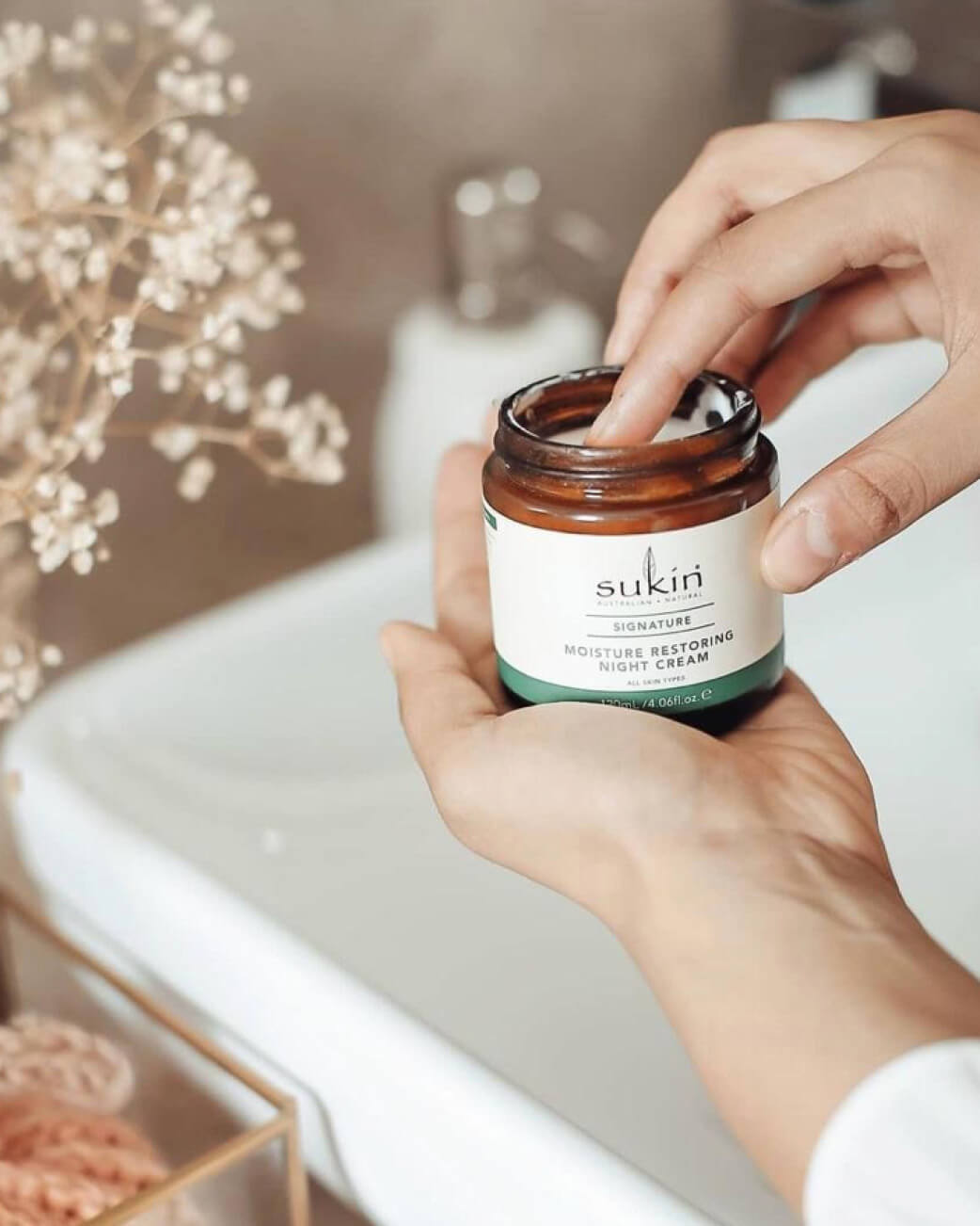 Hand holding a jar of Sukin night cream with a blurred bathroom background.