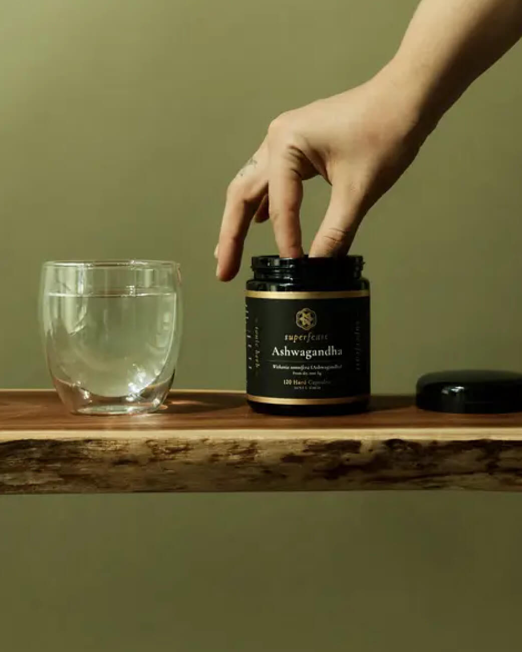 Hand opening a jar of Superfood Ashwagandha on a wooden surface with a glass of water.