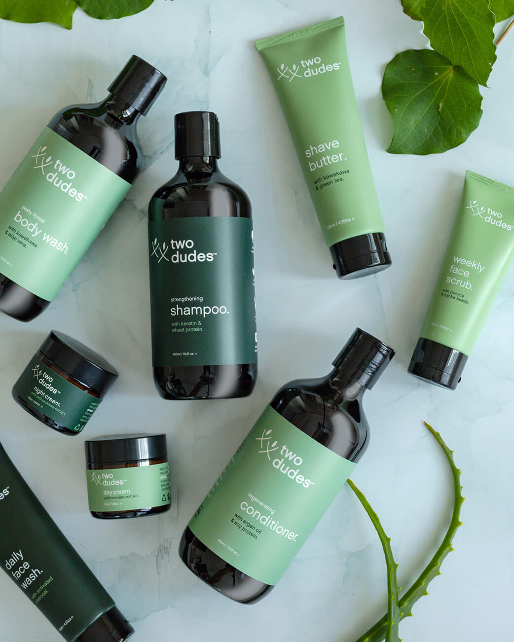 Collection of 'two dudes' skincare products on a white background with green leaves.