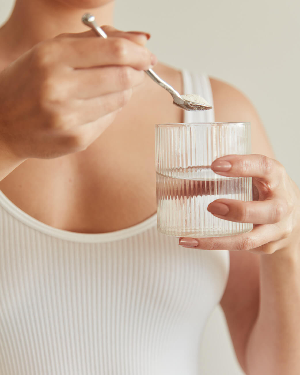 Person holding a glass of water and a spoon with a white powder, possibly collagen, against a neutral background.