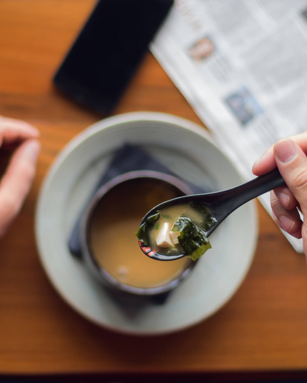 Spoon holding miso soup with seaweed over a bowl on a wooden table.
