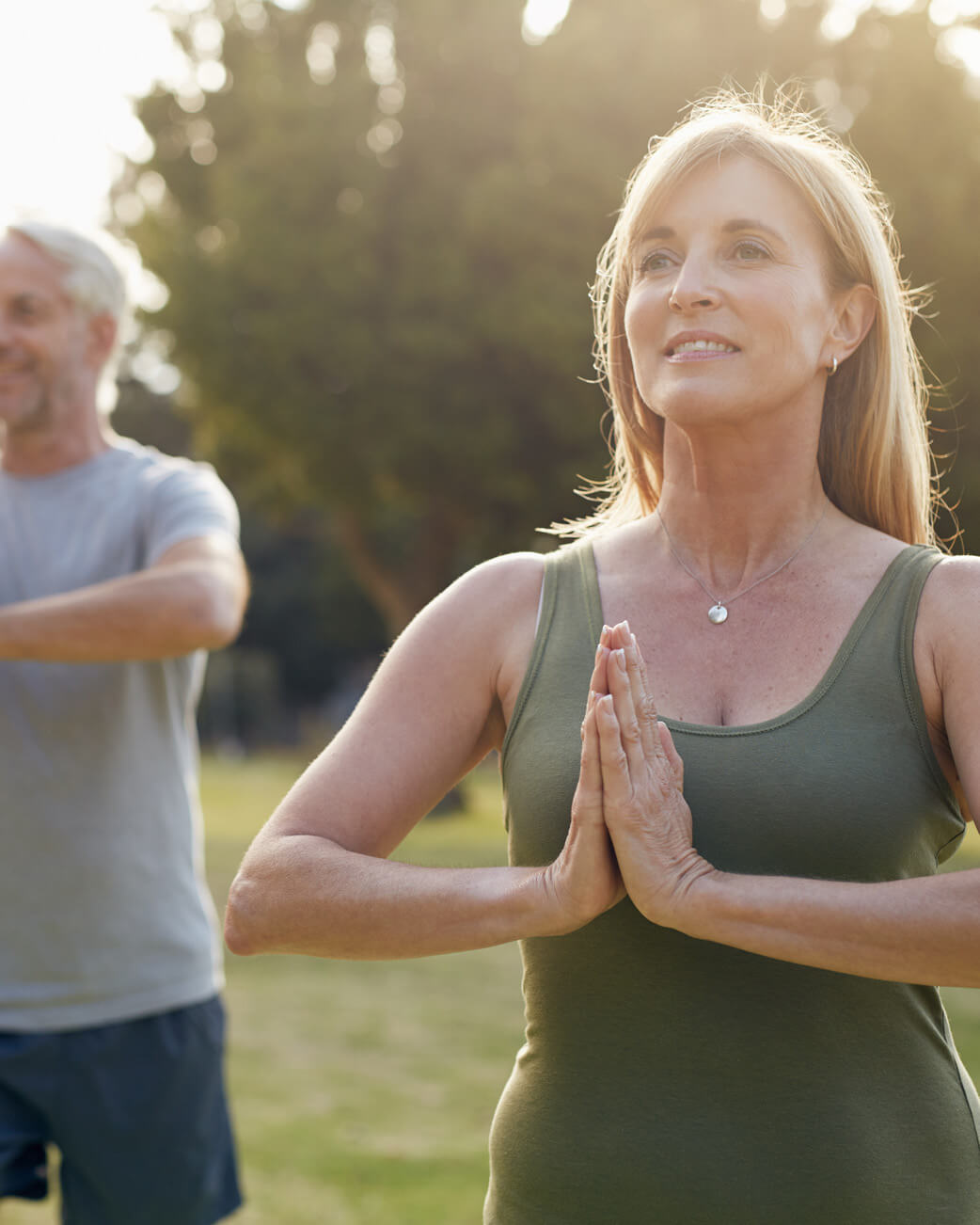 Woman in green tank top practicing yoga outdoors with a man in the background.