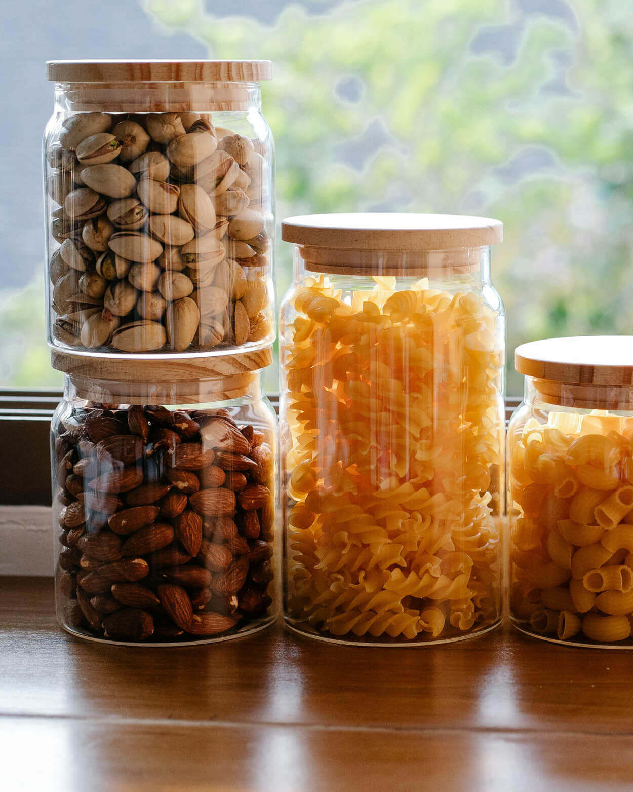 Four glass jars with wooden lids containing various dry foods on a wooden surface.