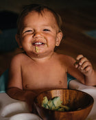 Baby eating from a wooden bowl with food on a dark background.