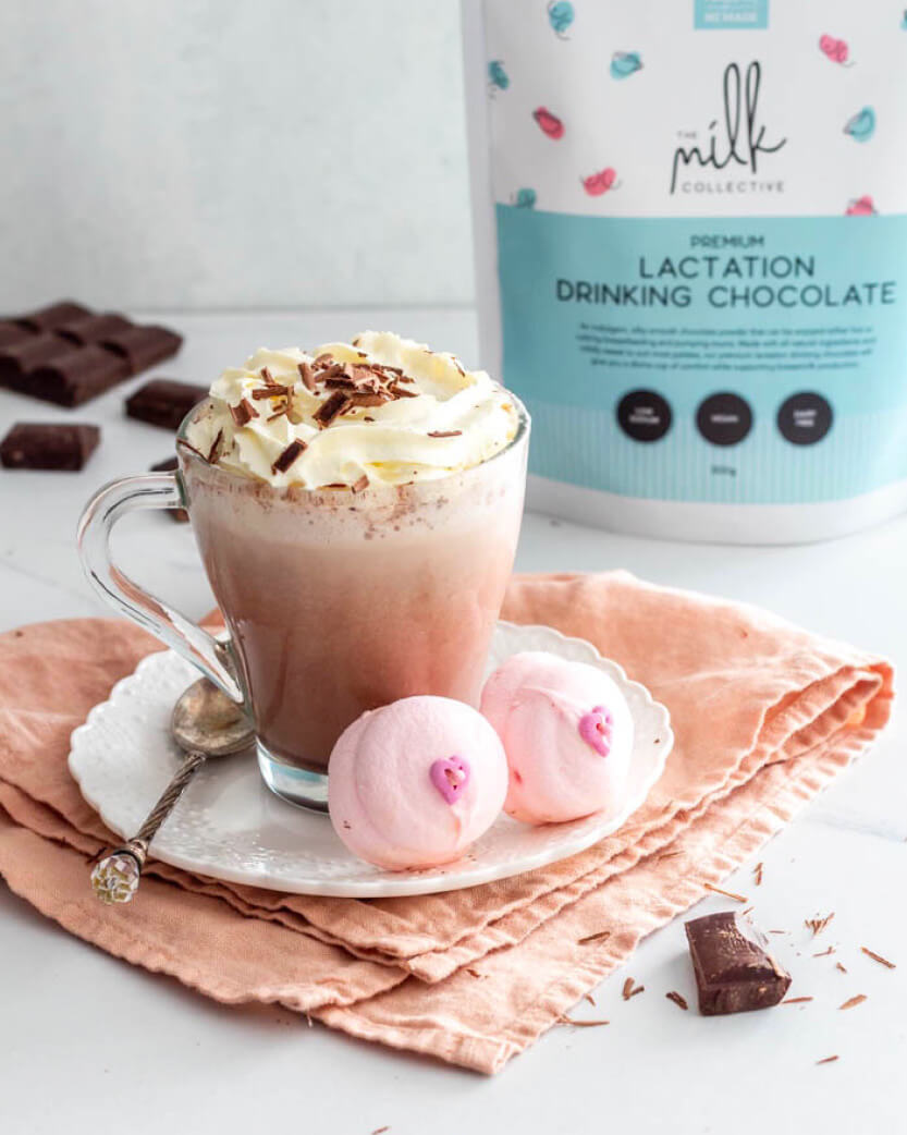 Hot chocolate with whipped cream & pink marshmallows on a white surface, with a bag of lactation drinking chocolate in the background.