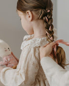 Child with braided hair being styled by an adult, wearing a lace-trimmed dress.