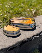 Two metal lunch containers with food on a stone surface outdoors.