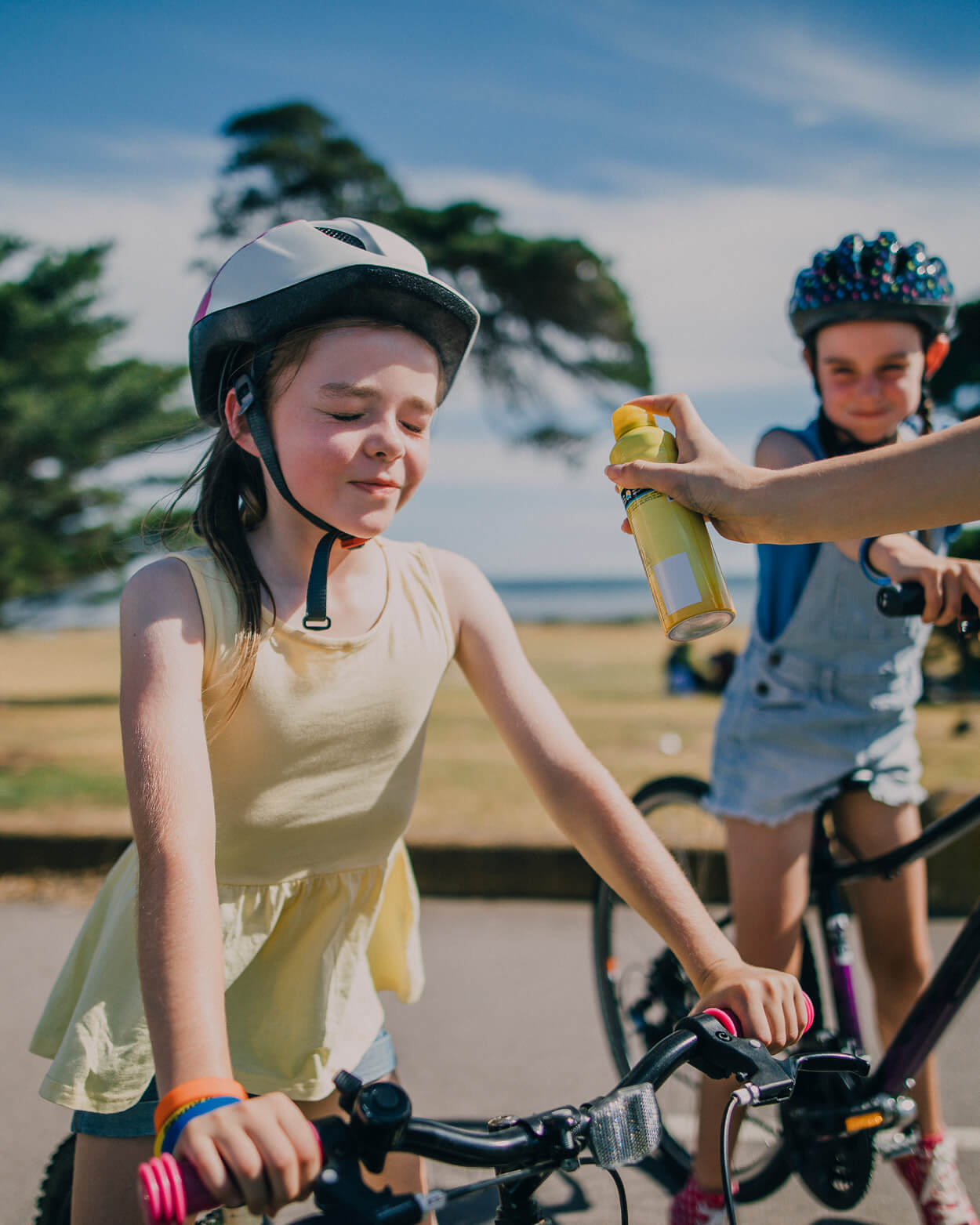 Two children on bicycles with helmets, one being sprayed with sunscreen.