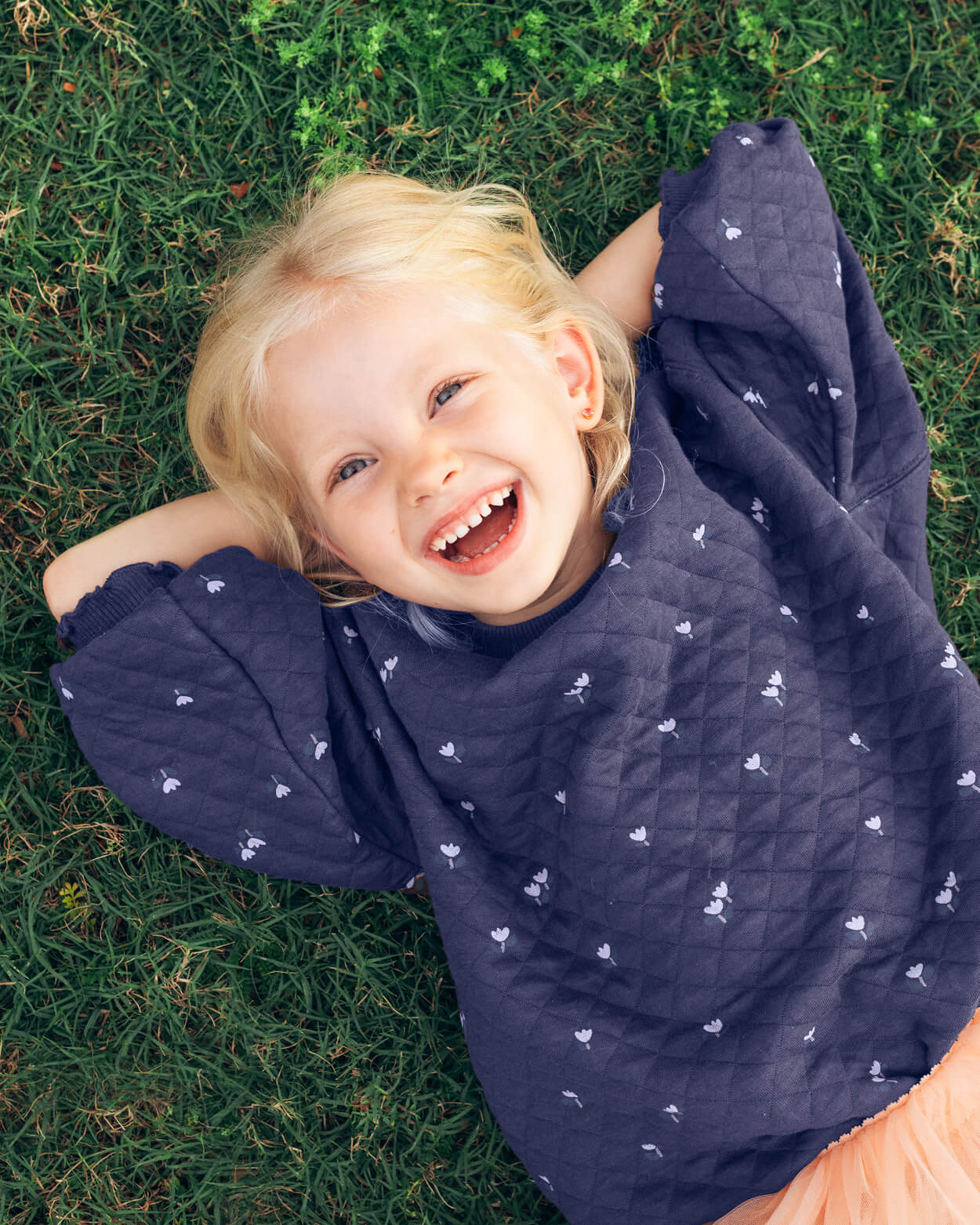Child wearing a navy blue quilted jacket with white patterns, lying on grass and smiling.