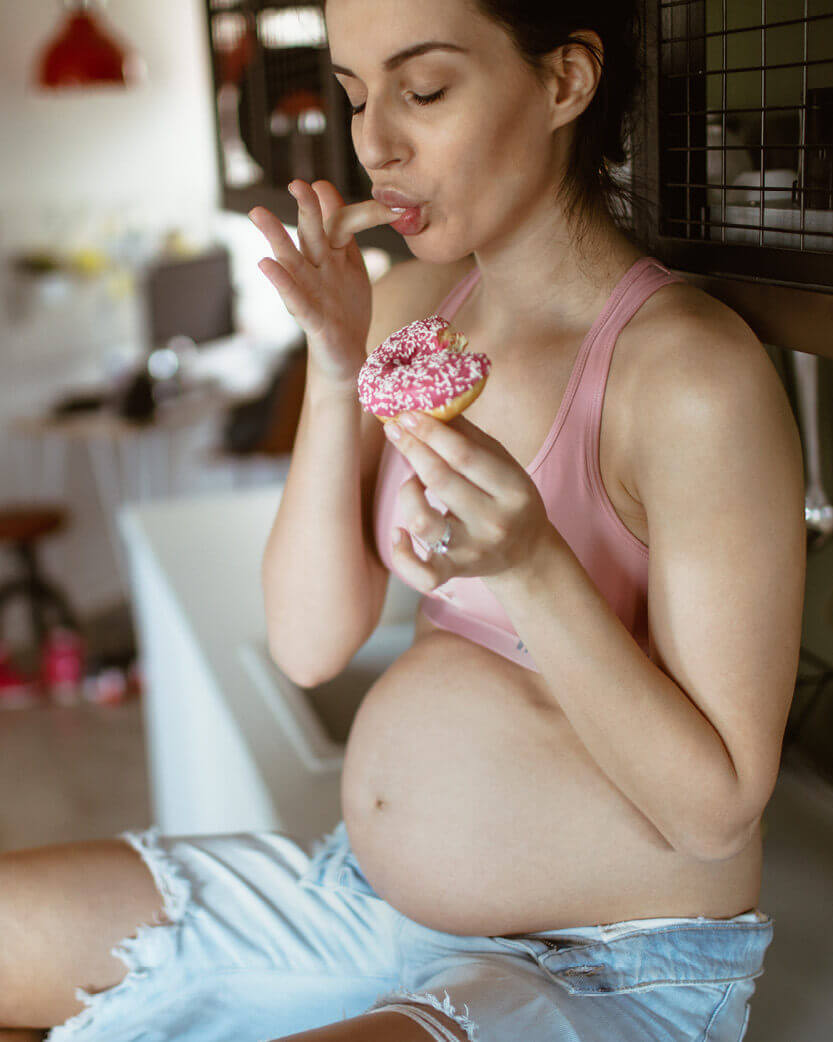 Pregnant woman eating a pink frosted donut in a casual setting.