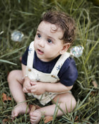 Child sitting in grass wearing a navy shirt and beige overalls.