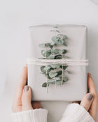 Gift wrapped in white paper with eucalyptus leaves, held by hands against a light background.