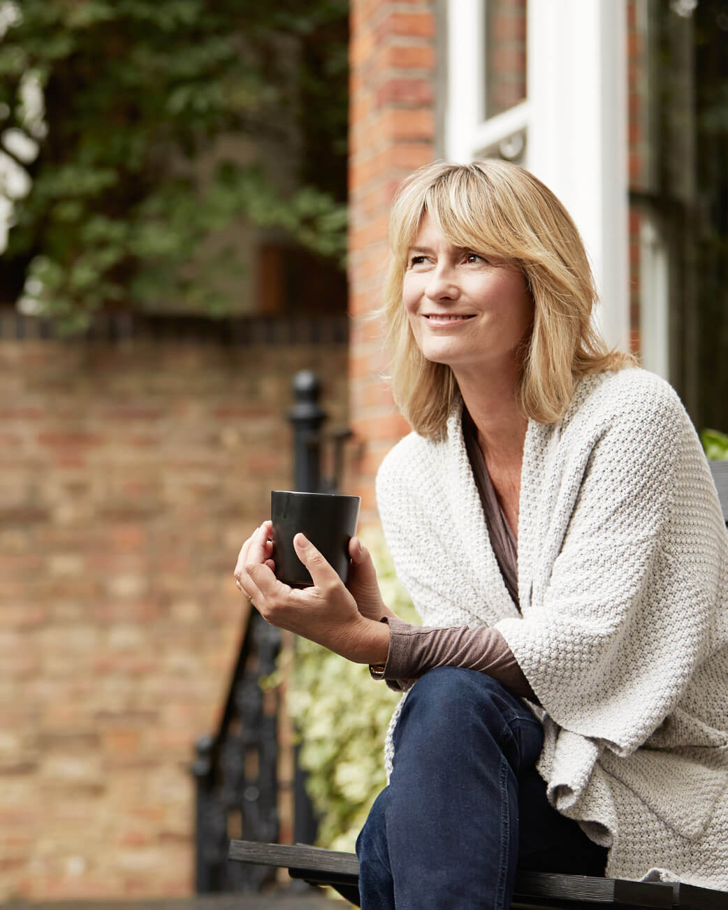 Woman sitting outdoors holding a cup, wearing a light-colored cardigan.