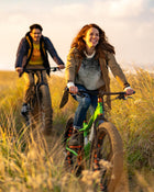 Two people riding bicycles through tall grass at sunset.