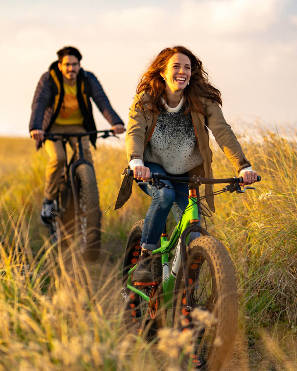 Two people riding bicycles through tall grass at sunset.