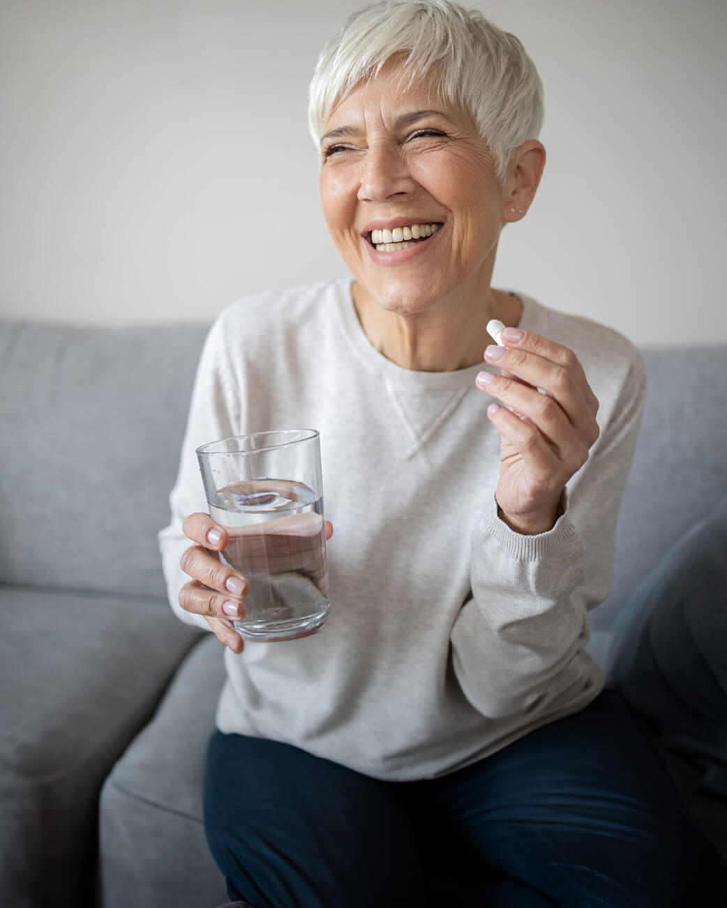 Woman holding a glass of water and a pill, sitting on a couch.