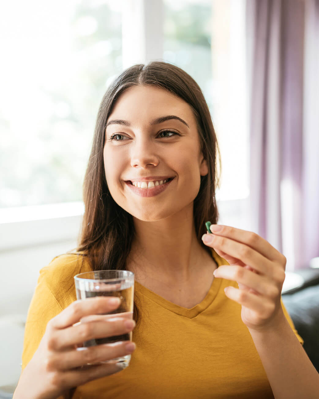 Woman holding a glass of water and a supplement pill indoors.