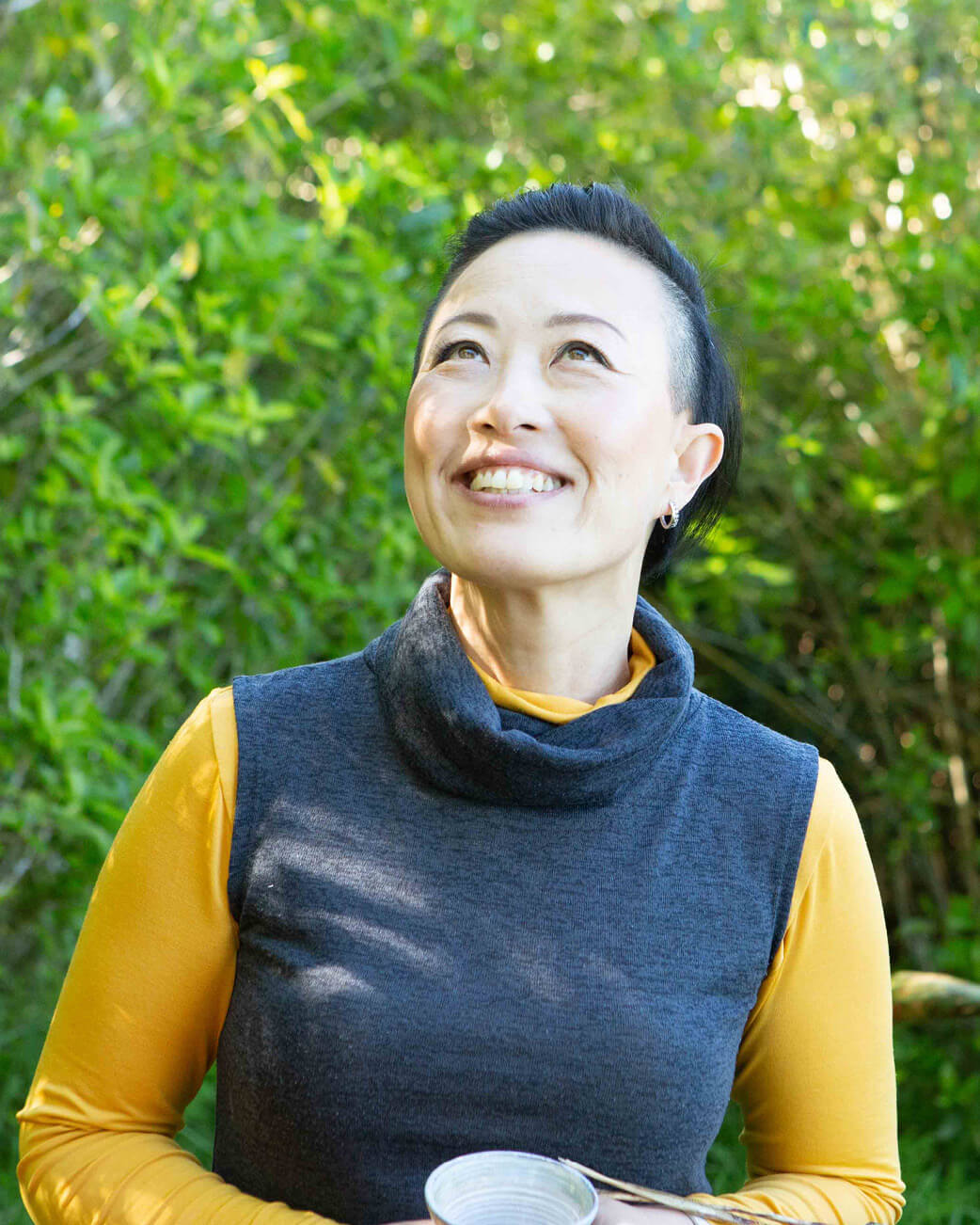Woman wearing a blue vest over a yellow shirt, standing outdoors with greenery in the background.