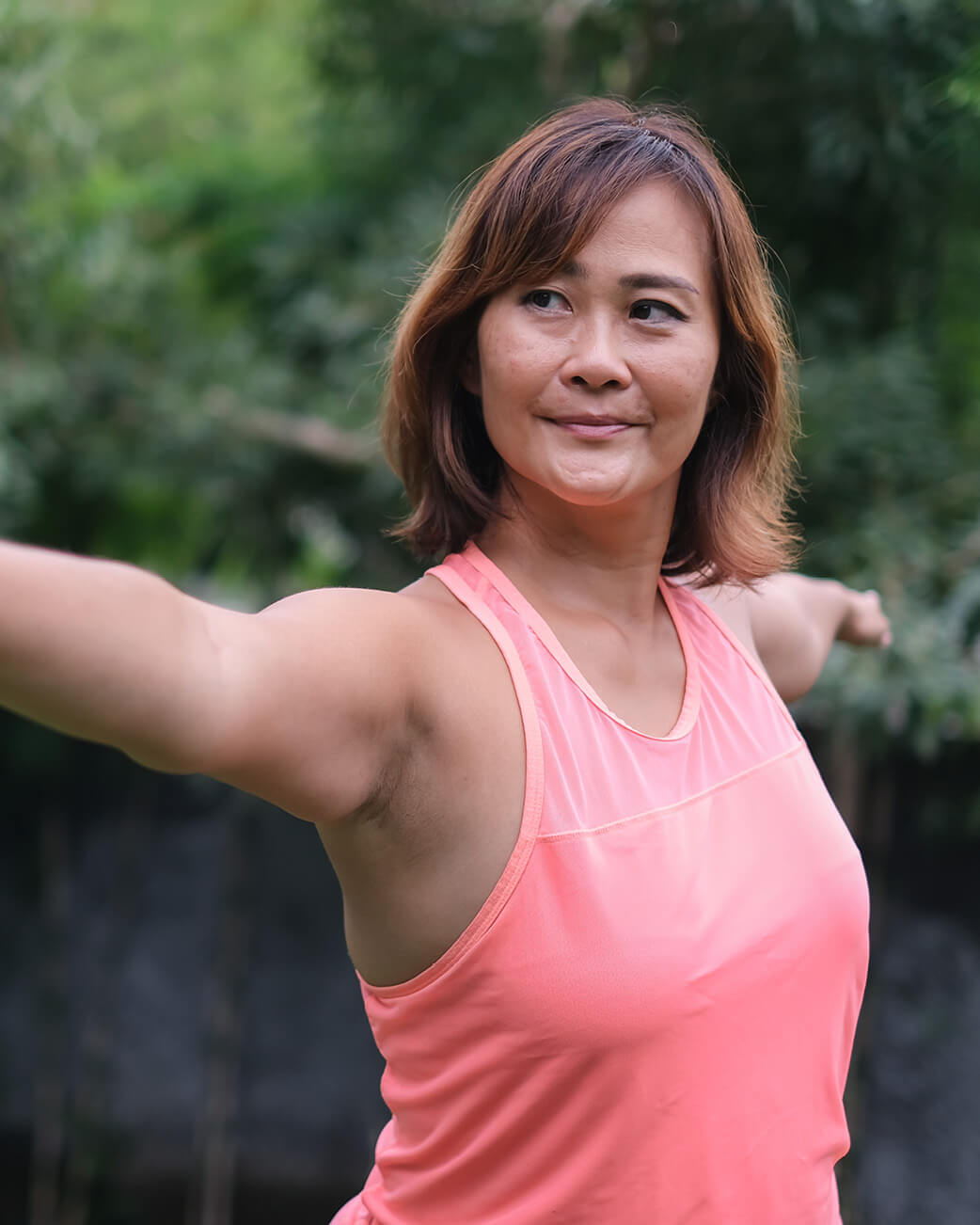 Woman in a pink tank top standing outdoors with greenery in the background.