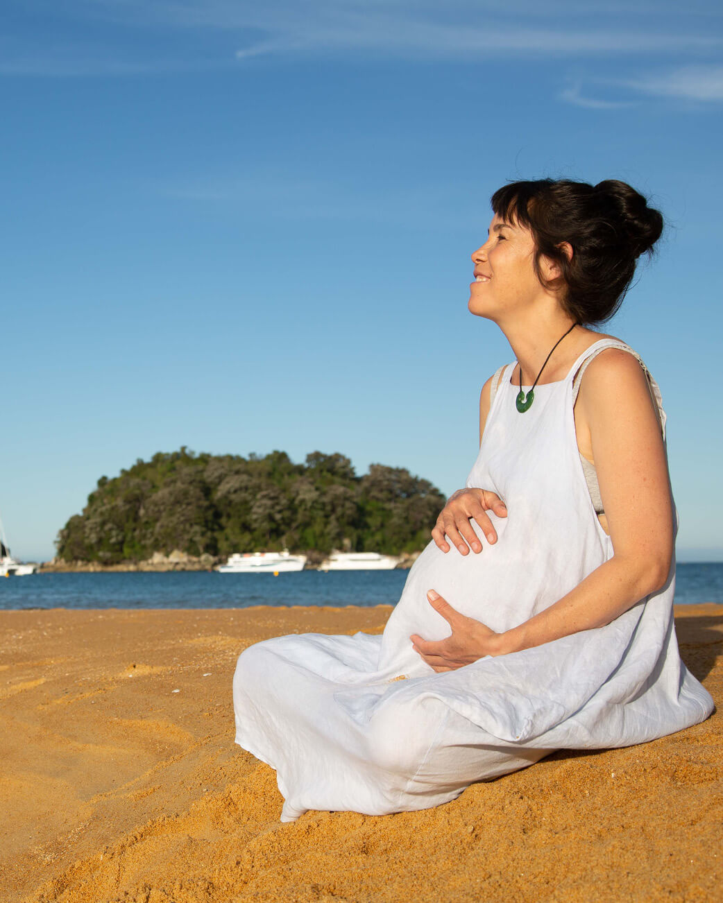 Pregnant woman in a white dress sitting on a sandy beach with a clear blue sky and water in the background.