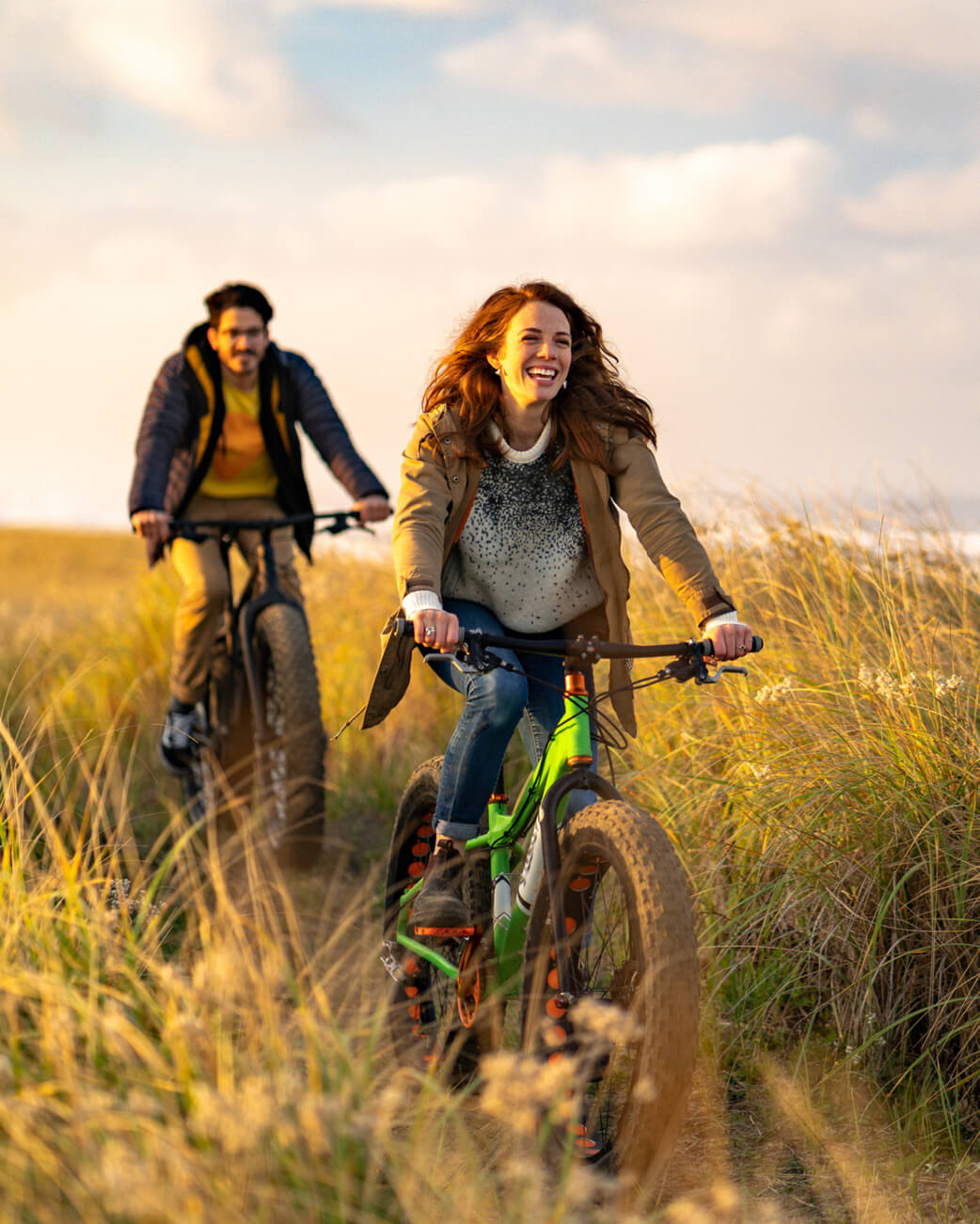 Two people riding bicycles through a grassy field with a scenic background.