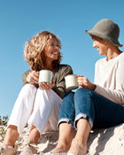 Two people sitting on a beach, holding mugs and smiling at each other.