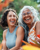 Two women laughing together outdoors with a blurred natural background.