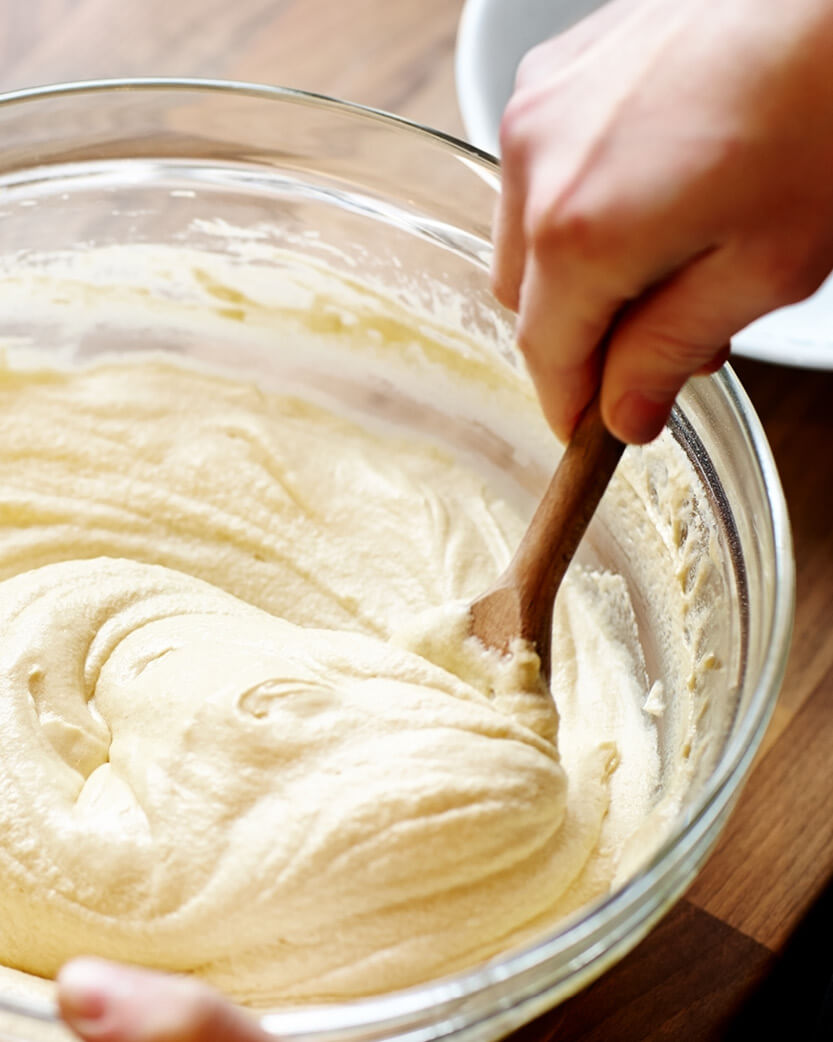 Person stirring creamy mixture in a glass bowl with a wooden spoon on a wooden surface.