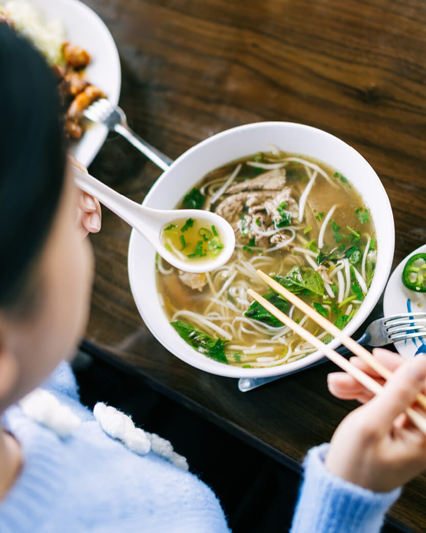 Person eating noodles with chopsticks from a bowl on a wooden table