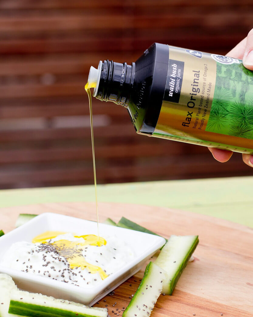 Person pouring flaxseed oil from a bottle onto a dish with vegetables on a wooden surface.