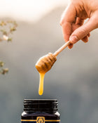 Hand holding a honey dipper with honey dripping from it, against a blurred natural background.